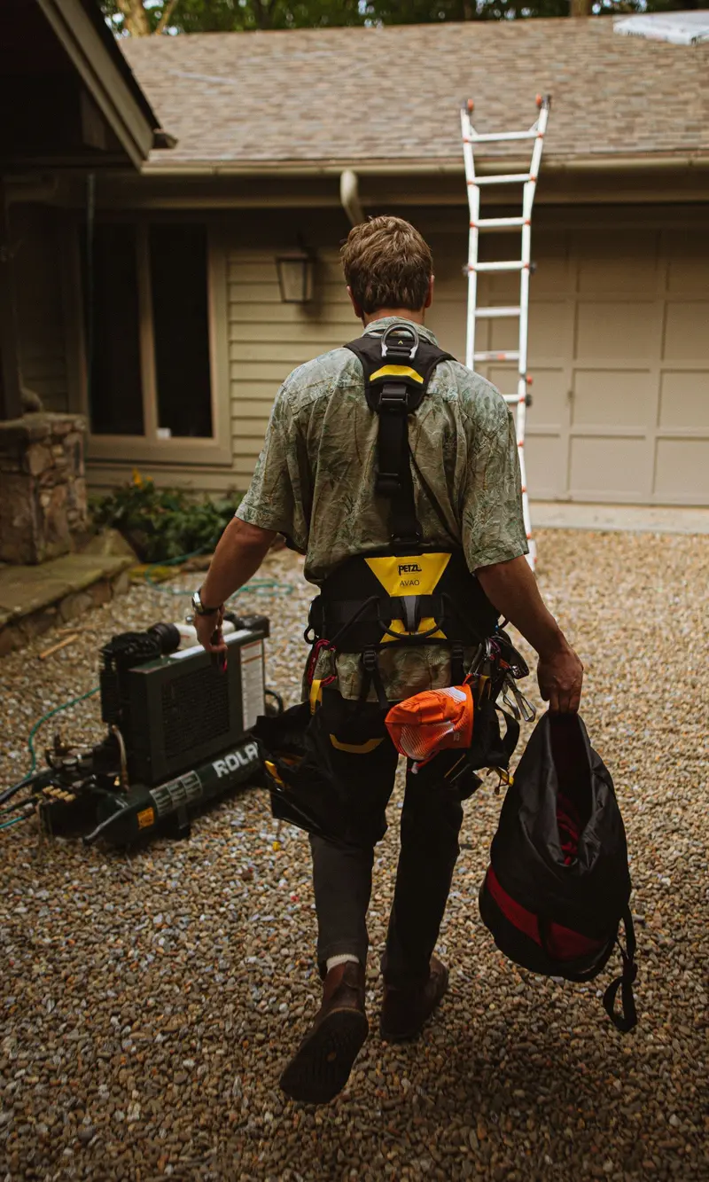 Roofer with a harness carrying gear toward a ladder at a house
