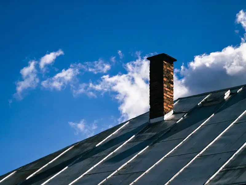 Dark standing-seam metal roof with a brick chimney against a blue sky